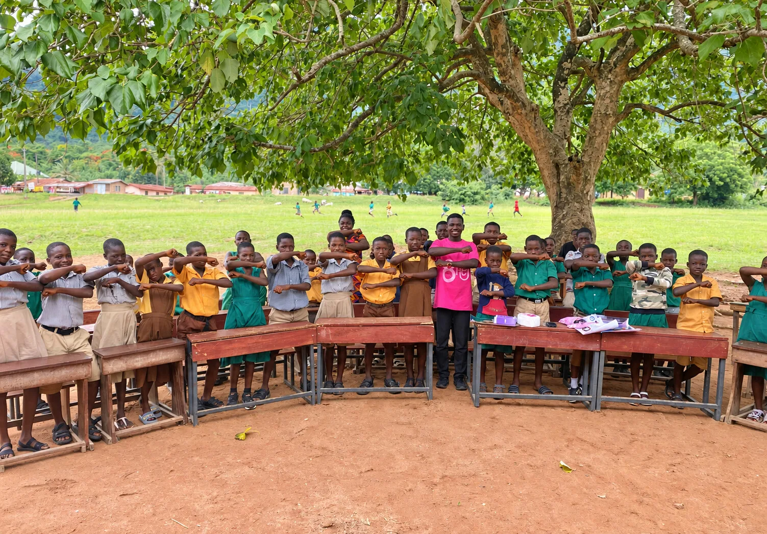 Schoolchildren and a SMART Hub facilitator standing behind desks under a tree during a Reach for Her activity.